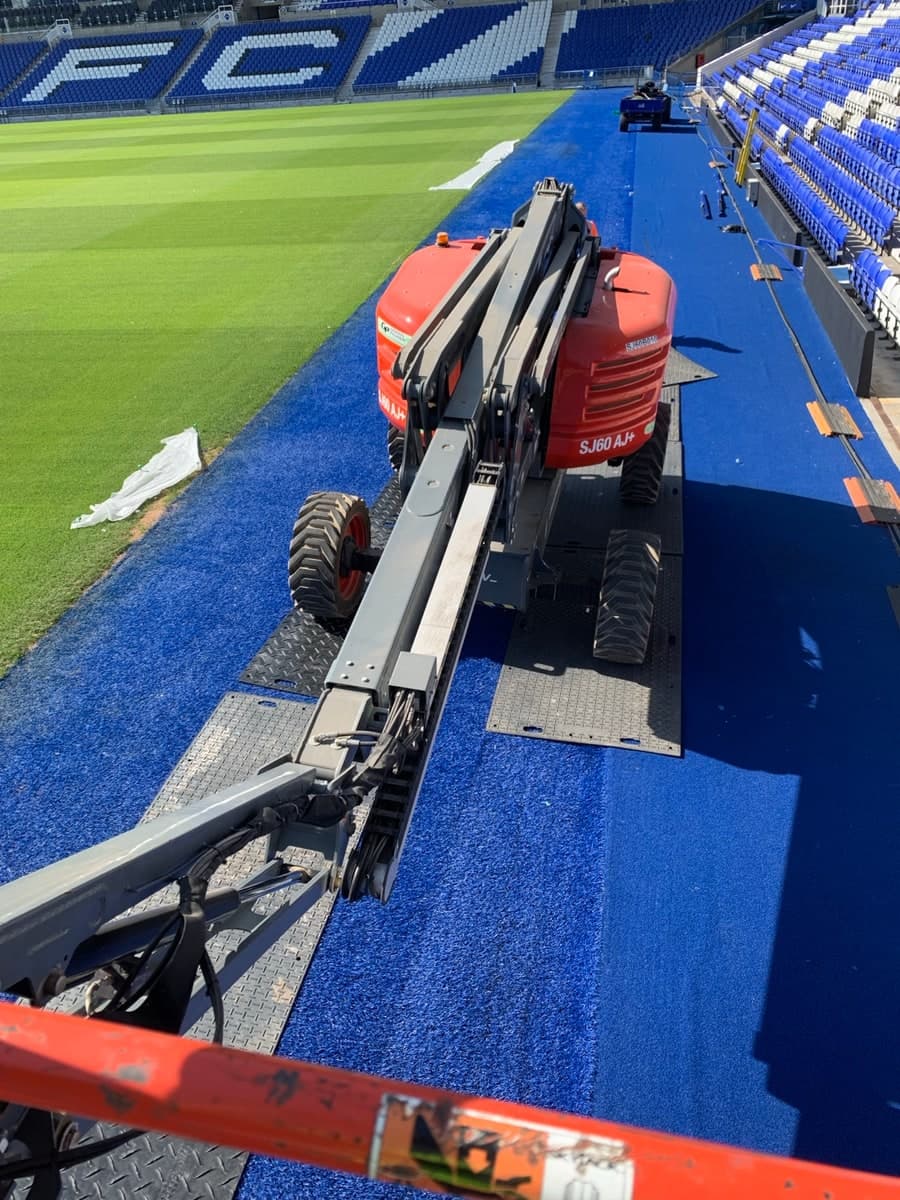 Boom lift positioned on protective matting beside a football pitch