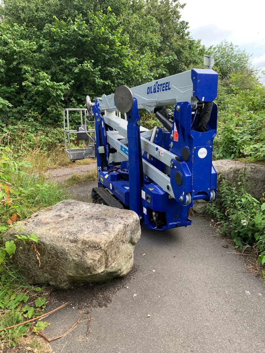 Folded tracked spider lift on a narrow path beside a large boulder in Levenshulme