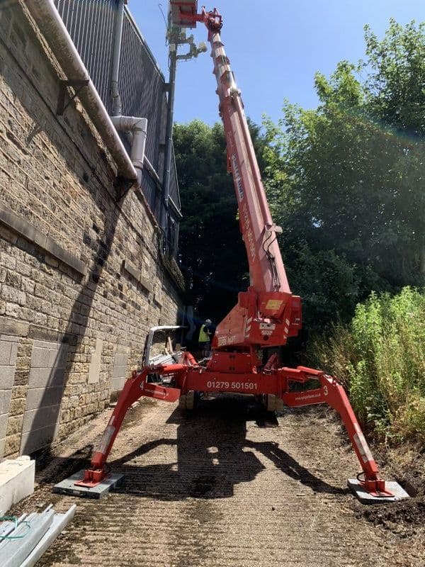 Tracked spider lift deployed in a tight-access corridor between stone walls, outriggers extended